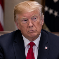 Donald Trump sits at a formal meeting table in a dark suit and red tie, with US flags blurred behind him.