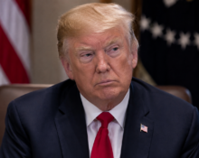 Donald Trump sits at a formal meeting table in a dark suit and red tie, with US flags blurred behind him.