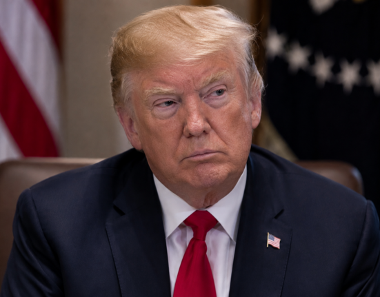 Donald Trump sits at a formal meeting table in a dark suit and red tie, with US flags blurred behind him.