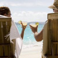 people sit in two wooden beach chairs facing the Caribbean ocean holding drinks and making a toast