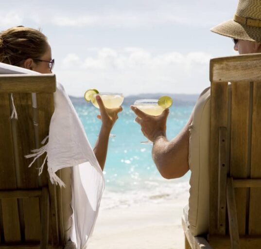 people sit in two wooden beach chairs facing the Caribbean ocean holding drinks and making a toast