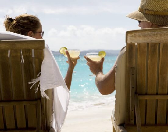 people sit in two wooden beach chairs facing the Caribbean ocean holding drinks and making a toast